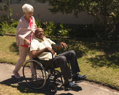 An elderly woman pushing an older man in a wheelchair along a garden path, both smiling and enjoying a sunny day outdoors.