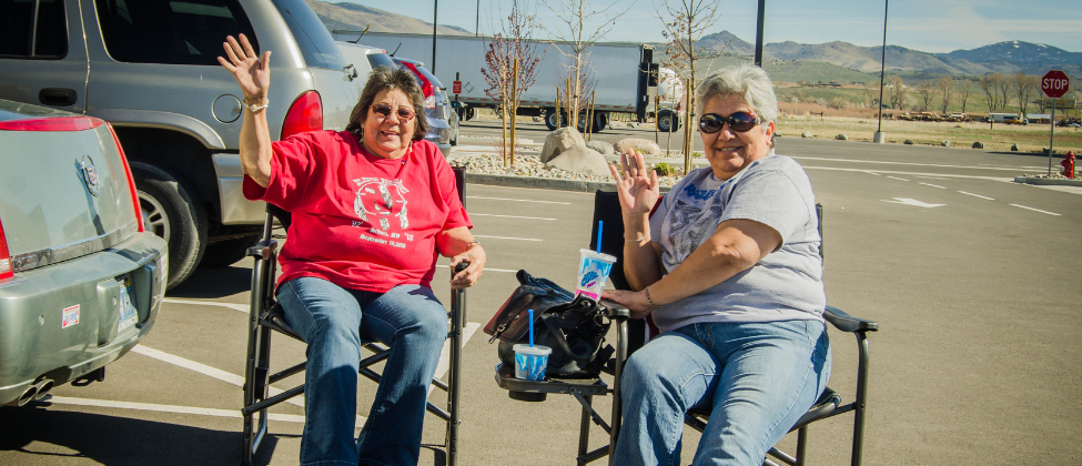 Two women sitting outside in a parking lot, smiling and waving while enjoying drinks in a rural community setting, symbolizing connection and support in underserved areas.