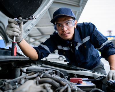 Focused auto mechanic in blue uniform inspecting and repairing a car engine, using a screwdriver in a well-equipped garage.