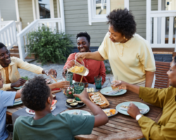 A joyful family gathering around a wooden dining table outdoors, with a woman pouring a drink while others engage in lively conversation, enjoying a meal together on a sunny day