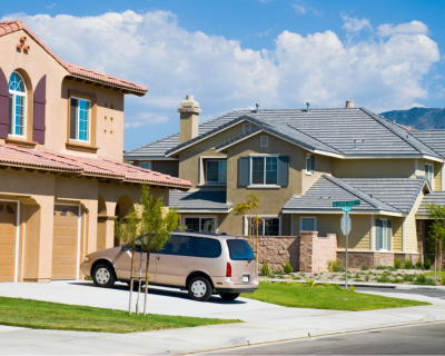 A picturesque suburban neighborhood featuring two modern houses with neatly maintained lawns and a parked minivan in a driveway under a sunny, blue sky.