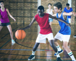 Two young athletes playing basketball in a gym, with a boy in a red jersey dribbling past a defender in a blue jersey, while other players are visible in the background.