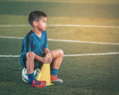 Young boy in a blue soccer jersey sitting on a yellow cone on a lush green soccer field, looking thoughtfully towards the horizon during sunset.